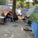 Several tents make up an encampment. In the background, a tree. Slightly left of center, an ETS outreach worker kneels in front of a client as they talk together.