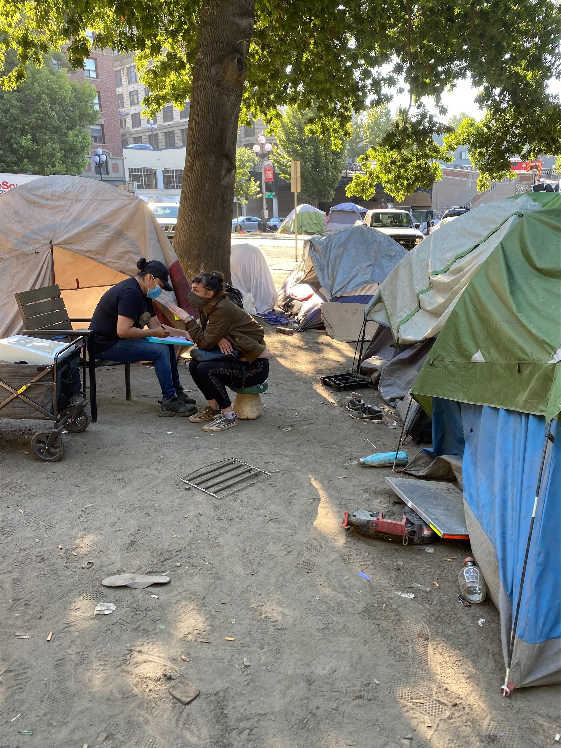 Several tents make up an encampment. In the background, a tree. Slightly left of center, an ETS outreach worker kneels in front of a client as they talk together.