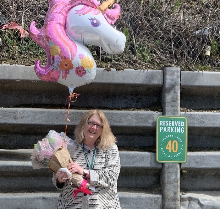 carol davidson, a white woman with shoulder length blond hair, stands beside a green parking plaque the denotes a reserved spot for an employee who has worked with ETS for 40 years. Carol is smiling and holding a bouquet of flowers and a large balloon shaped like a unicorn.