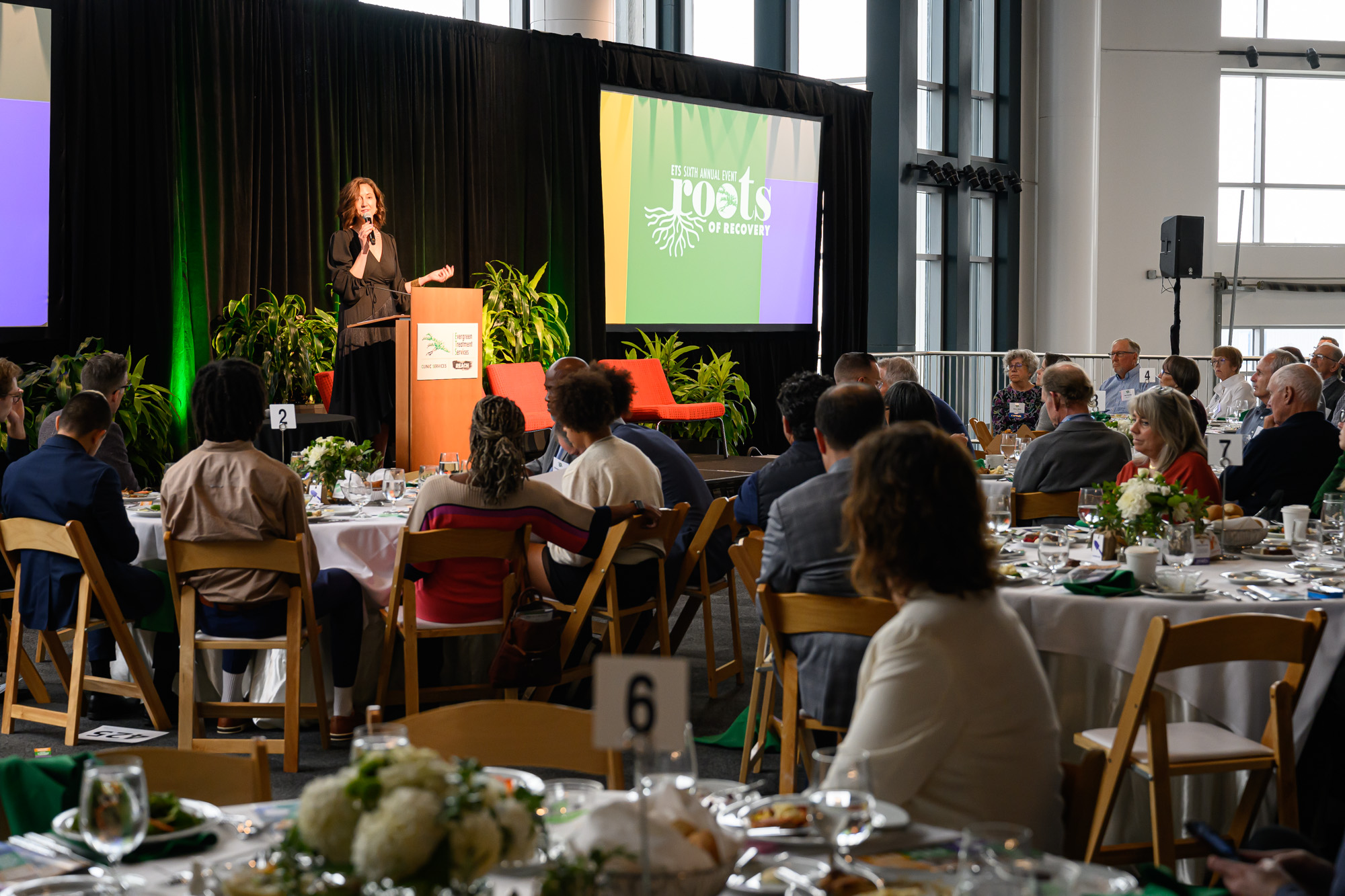 A white woman stands at a podium on stage where the screen behind her reads Roots of Recovery. An audience at round tables watch her speak.