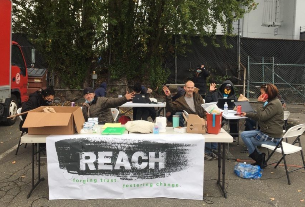 A white table sits outdoors with a white banner in front of it that reads REACH. There are people sitting around the table waving.